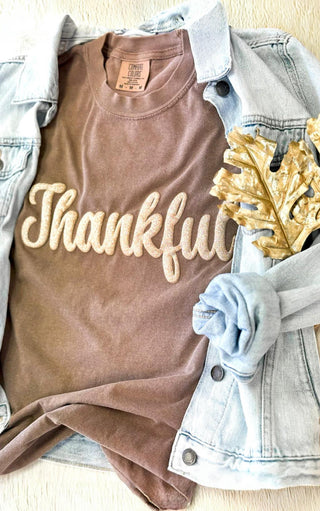 Close-up flat lay of brown Comfort Colors “Thankful” tee with glitter puff print, paired with a light denim jacket and gold leaf accent on a cream background.