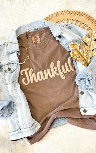 Brown Comfort Colors “Thankful” graphic tee with glitter puff print, styled flat lay with light denim jacket, gold leaf accent, and pearl bracelet on a neutral background.