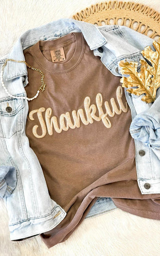 Brown t-shirt with 'Thankful' puffed print text, paired with a denim jacket on a light wooden surface.