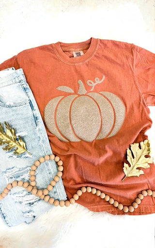 Close up view of a rust-colored Comfort Colors tee with a glitter puff print pumpkin design, styled flat lay with distressed light-wash jeans, gold leaf accents, and wooden bead garland on a cream background.