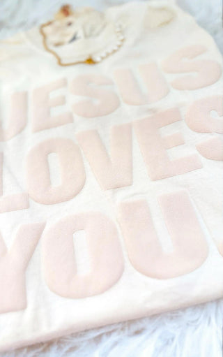 Close-up view of ivory Jesus Loves You puff print tee showing raised tone-on-tone lettering, styled with pearl and gold necklace on a white textured background.