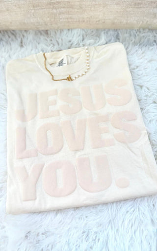 Ivory Jesus Loves You puff print tee folded on a white textured surface, styled with a delicate pearl and gold necklace.