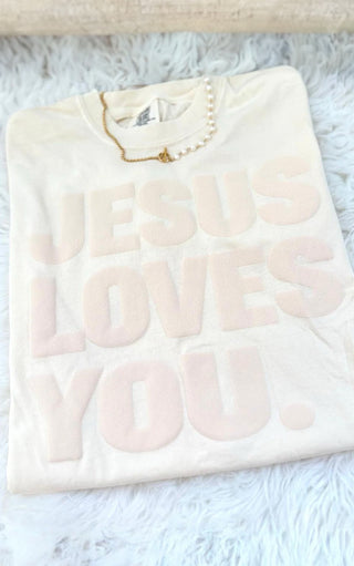 Ivory Jesus Loves You puff print tee folded on a white textured surface, styled with a delicate pearl and gold necklace for a chic neutral flat lay.