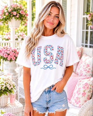 Blonde model standing on a sunny porch wearing a white patriotic USA tee with floral appliqué letters, embroidered blue bow, and distressed denim shorts.