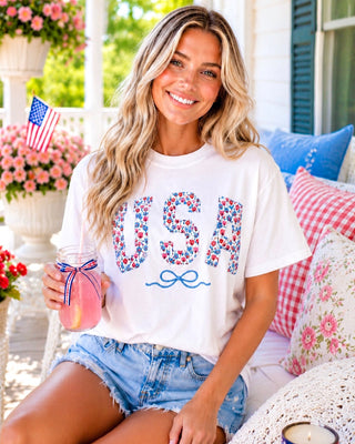 Blonde model posing on a sunny porch wearing a white patriotic USA tee with floral appliqué letters, embroidered blue bow, and distressed denim shorts.