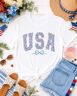 Flat lay of White USA t-shirt featuring floral tulip patch letters and embroidered blue bow styled with denim shorts, sandals, and patriotic summer accessories.