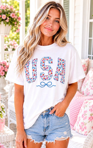 Smiling blonde model wearing a white USA t-shirt with floral tulip patch letters and an embroidered blue bow, styled with distressed denim shorts on a Southern front porch.