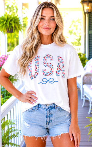 Woman wearing a white t-shirt with 'USA' and a bow graphic, standing outdoors.