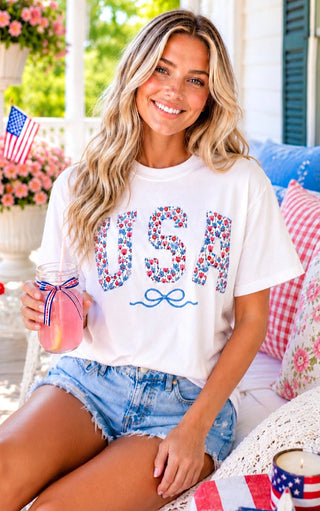 Smiling blonde model wearing a white USA t-shirt with floral patch letters and an embroidered blue bow, styled with distressed denim shorts on a Southern front porch.