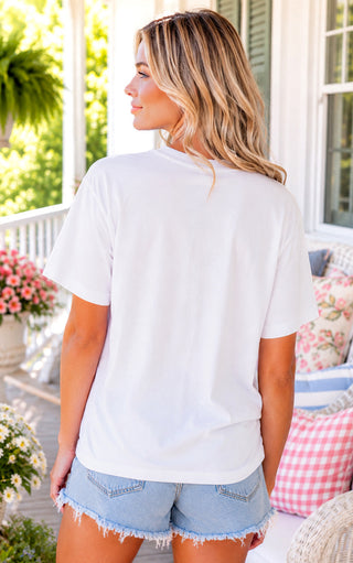 Back view of Woman wearing a white t-shirt and denim shorts on a porch with flowers and furniture. 