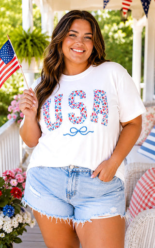Smiling curvy model holding an American flag while wearing a white floral USA shirt with embroidered blue bow and denim shorts.