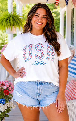 Curvy brunette model wearing a white USA t-shirt with floral tulip patch letters and embroidered blue bow, styled with distressed denim shorts.