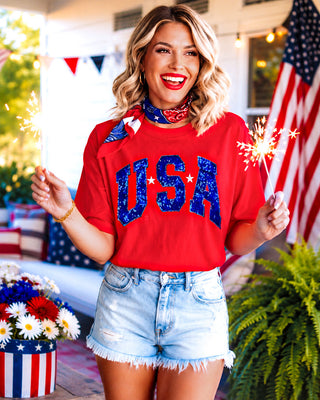 Woman wearing a red 'USA' shirt with sparklers and an American flag in the background