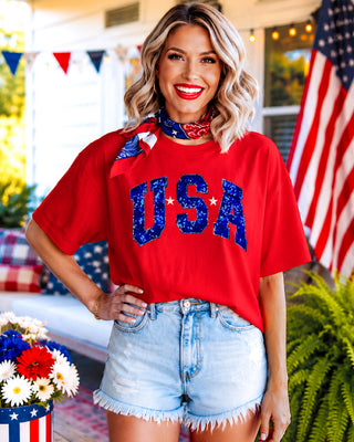 Woman wearing a red 'USA' shirt with blue shorts in a patriotic setting.