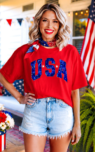 Woman wearing a red 'USA' t-shirt with denim shorts in an outdoor setting.