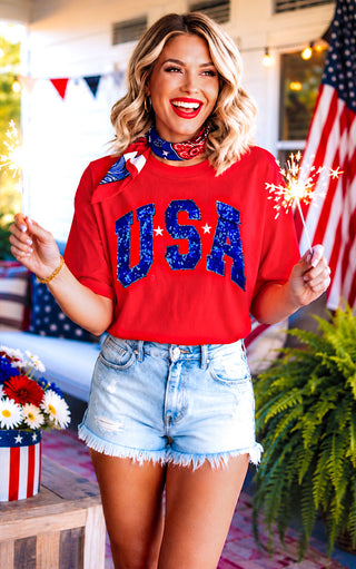Woman wearing a red 'USA' shirt with blue sequins, holding a sparkler outdoors.