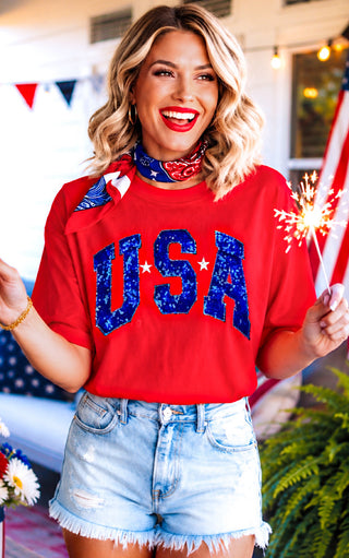 Woman wearing a red and blue sequin  'USA' shirt with blue shorts, holding a sparkler outdoors.