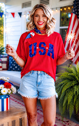 Woman wearing a red 'USA' shirt with sparklers on a decorated porch.