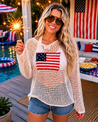 Woman wearing a white mesh top with an American flag design, holding a sparkler by a poolside.