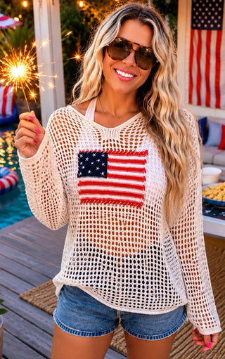 Woman wearing a white mesh top with an American flag design, holding a sparkler outdoors.