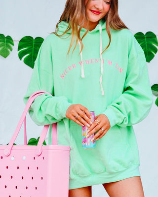 Person wearing a light green hoodie with text, holding a pink tote bag and colorful drink, against a light blue background with green leaves.