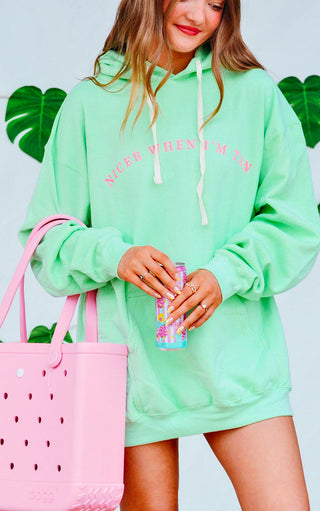 Person wearing a light green hoodie with text, holding a pink bag and colorful drink, against a light blue background with green leaf decorations.