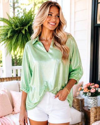 Woman wearing a seafoam green blouse and white shorts on a porch with plants and flowers.