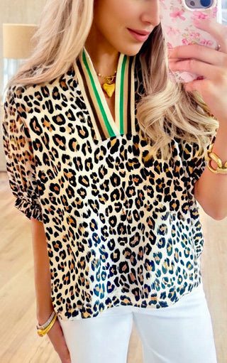 Mirror selfie of a woman wearing a leopard print blouse with striped trim split neckline and white pants, holding a phone with floral case.