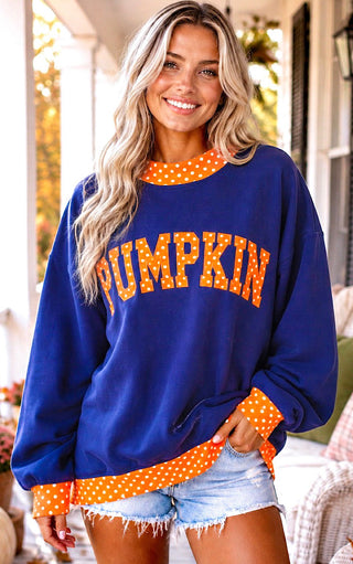 A woman wearing a royal blue crewneck sweatshirt with 'PUMPKIN' printed across the chest in orange polka-dot letters, featuring orange polka-dot trim on the collar, cuffs, and hem, paired with denim shorts.