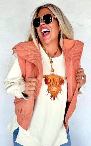 Woman smiling in a cream Highland Cow embroidered sweatshirt layered under a light rust vest, styled with casual jeans.