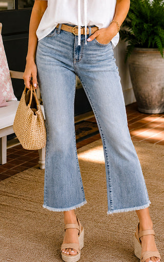 Close up of a woman wearing Judy Blue cropped kick flare jeans, a white blouse, and tan wedge sandals on a Southern front porch.