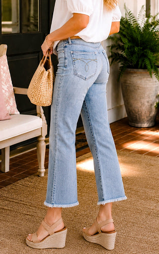 Close up side view of female  model wearing Judy Blue cropped kick flare jeans, a white  blouse, and tan wedge sandals on a Southern front porch.