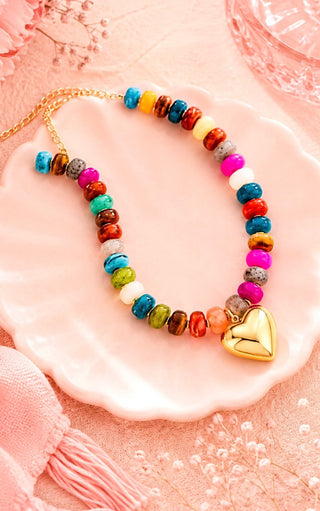 A colorful beaded necklace with a gold heart charm, displayed on a pink scalloped dish against a soft pink background.