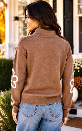 Back view of A woman wearing a brown mock neck knit sweater with white floral embroidery on the sleeves, shown from the back in an outdoor porch setting.