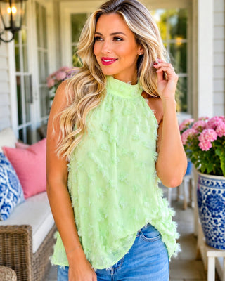 A woman wearing a sleeveless lime green textured top and blue jeans, standing on a porch with pink flowers and wicker furniture in the background.