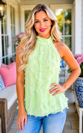 A woman modeling a sleeveless, high-neck lime green ruffled top with blue jeans, standing on a sunny porch with white columns and pink cushions in the background.