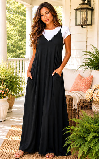 A woman stands on a porch wearing a black maxi slip dress with a white short-sleeve crewneck top underneath. The dress has spaghetti straps and side pockets, and she is wearing brown sandals.