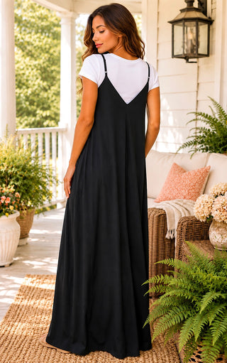 A woman stands on a porch wearing a long black slip-style maxi dress with thin straps and a white short-sleeve top layered underneath. The setting includes a wicker chair with pillows, potted ferns, and a white house exterior in the background.