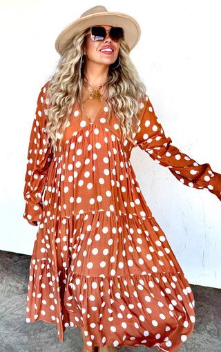 Model smiling in the rust polka dot dress with a wide-brim hat and sunglasses, sleeves slightly puffed at the cuffs.
