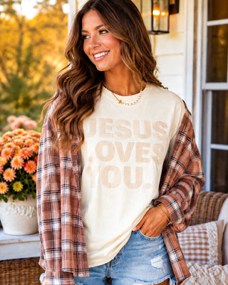 Woman wearing a 'Jesus Loves You' shirt with a plaid jacket on a porch.