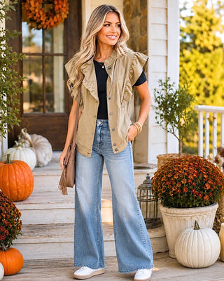 A woman stands on a decorated front porch wearing a sleeveless tan ruffle-trimmed vest over a black top, light blue wide-leg jeans, and white sneakers. Pumpkins, fall flowers, and a porch setting are visible around her.
