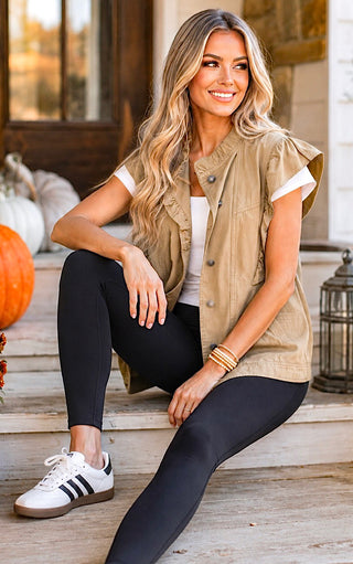 A woman sits on outdoor wooden steps wearing a sleeveless tan button-front vest over a white top, black leggings, and white sneakers with black stripes. Pumpkins and a lantern are visible in the background.