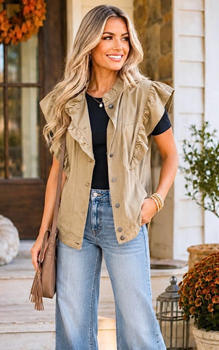 A woman stands on a porch wearing a sleeveless beige ruffle-trimmed button-front vest over a black top and light-wash blue jeans, holding a brown fringe shoulder bag.