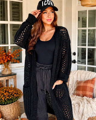 A woman stands on a porch wearing a long black open-knit cardigan over a black top and dark gray sweatpants, with a black baseball cap featuring the word 'ICON' in white. The background shows a home porch with white-framed glass doors, potted orange flowers, and a cream-colored chair.