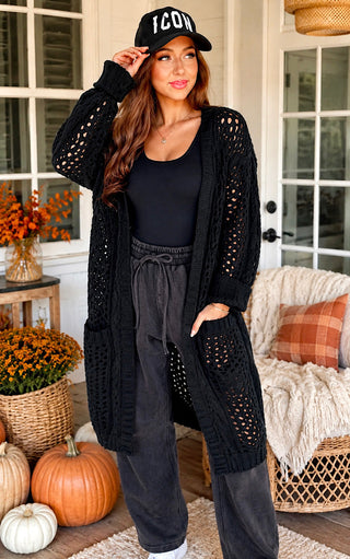 A woman stands on a porch wearing a long black open-knit cardigan over a black top and dark gray drawstring sweatpants, with a black baseball cap featuring white text. Pumpkins, flowers, and a woven chair are visible in the background.