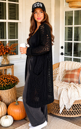 A woman stands on a porch wearing a black open-knit long cardigan over a black top and gray wide-leg pants, with a black cap that says ICON. She holds a white coffee cup.