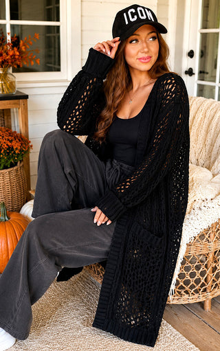 A woman sits on a porch wearing a long black open-knit cardigan over a black top and dark gray wide-leg pants, with a black baseball cap that says 'ICON' in white letters.