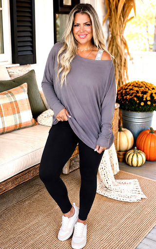 A woman modeling a loose, long-sleeve off-the-shoulder top in gray, paired with black leggings and white sneakers, standing on a decorated porch with pumpkins and autumn decor.