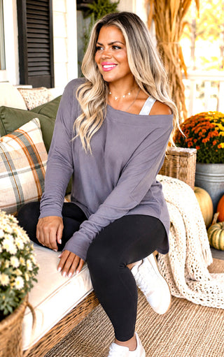 A woman sitting on a porch sofa wearing a loose, off-the-shoulder long-sleeve top in gray, black leggings, and white sneakers.