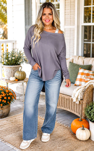 A woman stands on a decorated porch wearing a loose, off-the-shoulder long-sleeve top in muted gray  with wide-leg blue jeans and white sneakers.